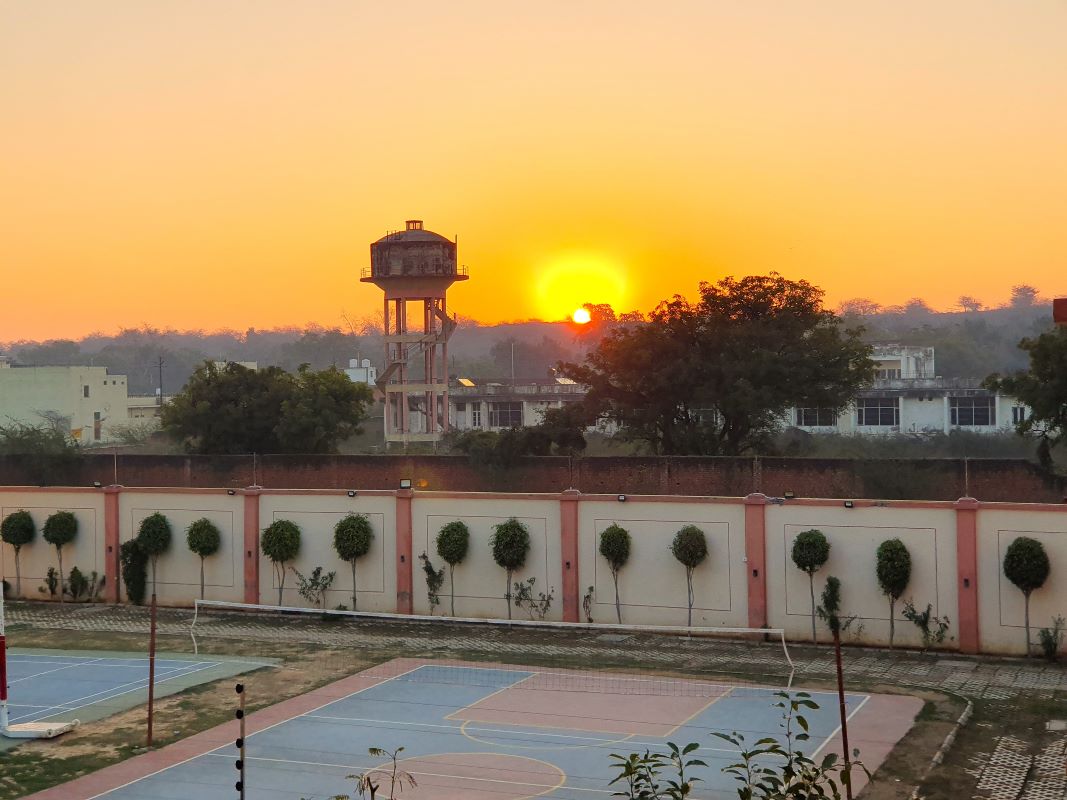 Februárban Indiában töltöttem két hetet. Reggelente mindig megcsodáltam a napkeltét az erkélyről. A háttérben a védikus írások által legszentebbnek tartott hegy, a Govardhan hegy látszik (ami valójában inkább egy domb.) A szomszédban egy iskola működik, annak a sportpályája látszik, a fal tövében levő fácskák pedig kislevelű fikuszok.